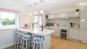 A modern kitchen with white cabinets, a central island with bar stools, and a view of the outdoors through a window. - Y Beudy (Caernarfon)