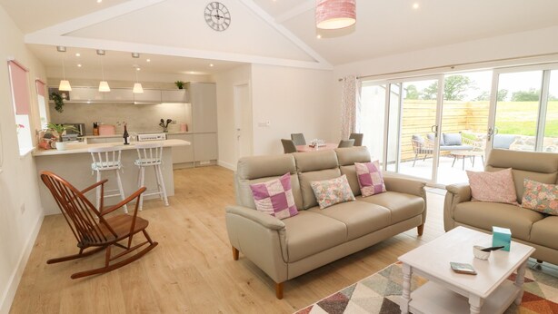 A spacious living area with a sofa, a rocking chair, and a coffee table. A kitchen is visible in the background with a dining area and large windows. - Y Beudy (Caernarfon)