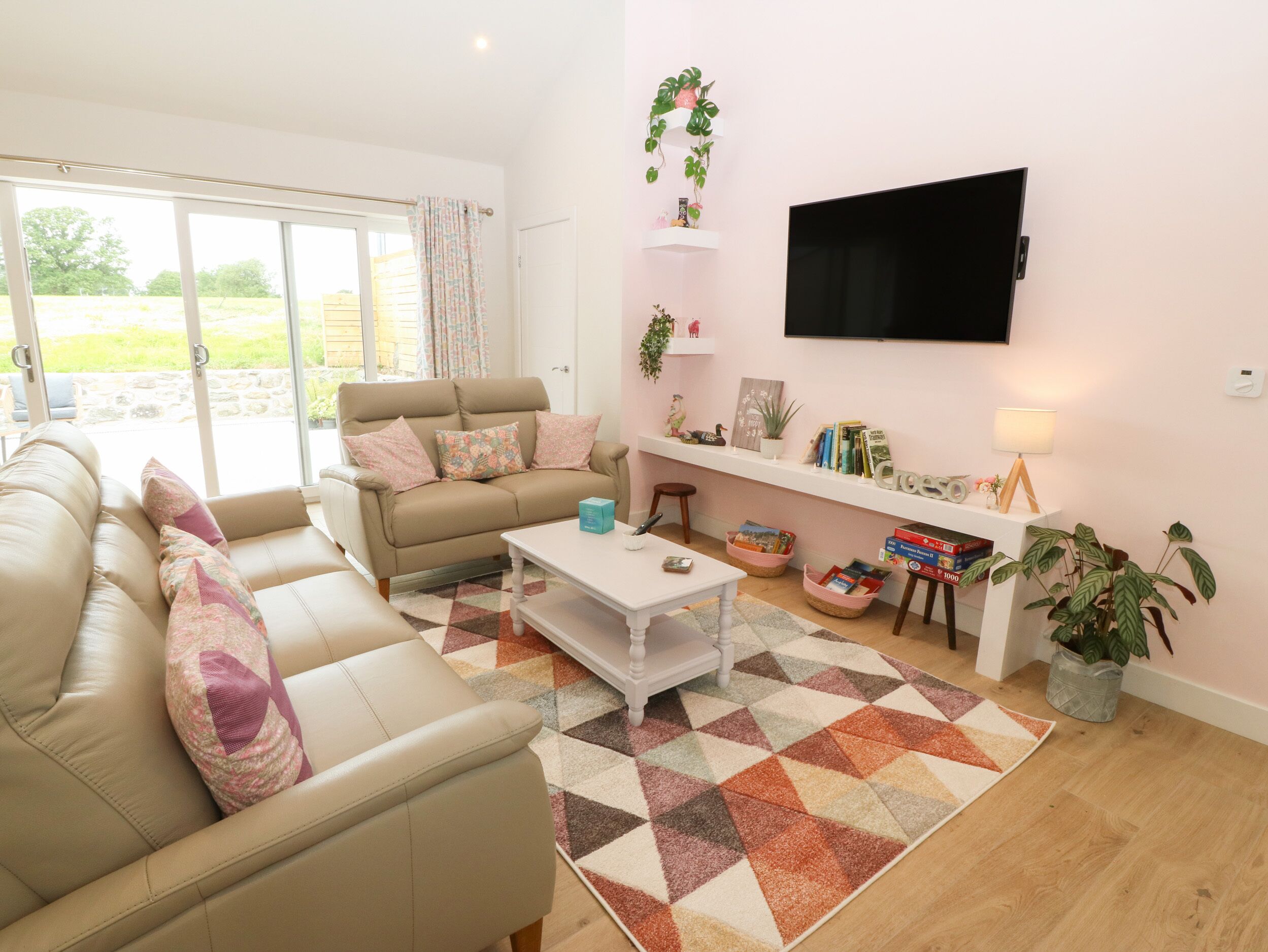 Una sala de estar con un sofá color beige, una mesa de centro, un televisor montado en la pared y una estantería con libros y objetos decorativos.