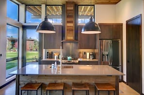A modern kitchen with a large island, two pendant lights, a stainless steel refrigerator, and a view of the outdoors. - Base Camp 49 - Flagg Mountain Cabin 7B (Mazama)