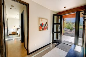 A hallway leading to a bedroom with a bed, a nightstand, and a lamp. There is a door leading outside with a view of the outdoors. - Base Camp 49 - Goat Peak Cabin 11A (Mazama)