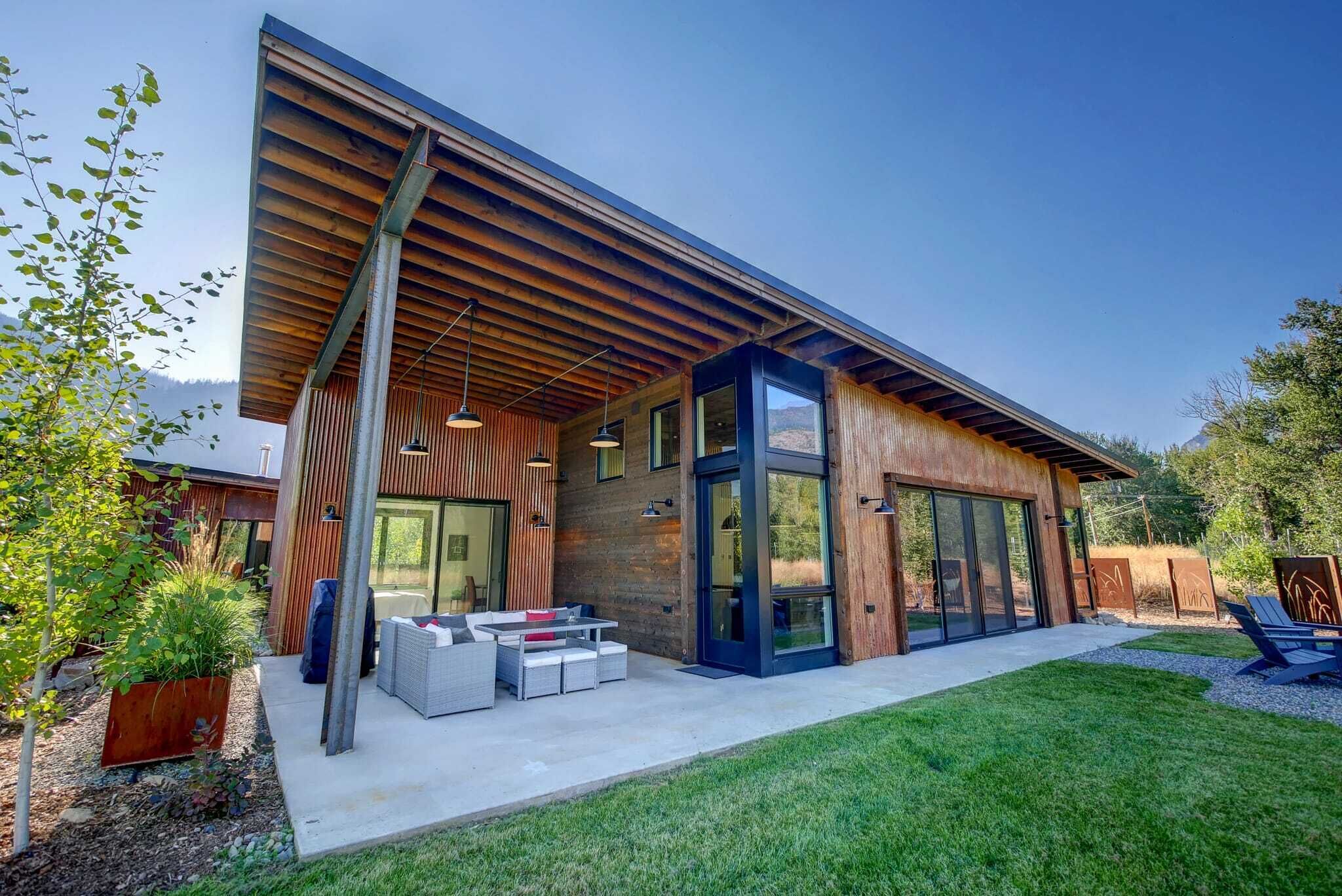 Une maison moderne avec un toit en pente, de grandes portes vitrées et un patio agrémenté de mobilier d’extérieur.
