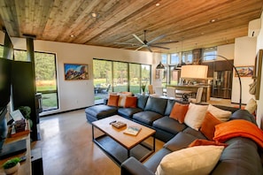 A modern living room with a large sectional sofa, a coffee table, and a TV. There is a kitchen area with a stove and refrigerator. The room has large windows and a ceiling fan.