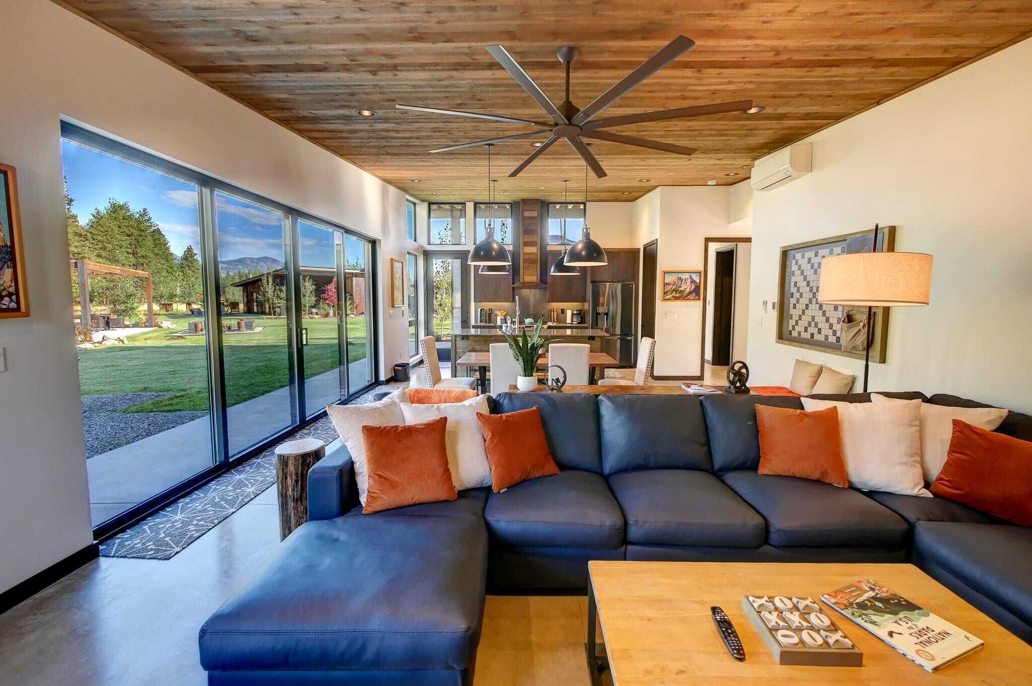 Un salon moderne avec un canapé d’angle, une table basse et une grande fenêtre donnant sur un jardin et des montagnes.