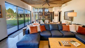 A modern living room with a sectional sofa, a coffee table, and a large window offering a view of a garden and mountains. - Base Camp 49 - Lucky Jim Cabin 11B (Mazama)