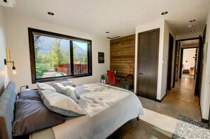 Un dormitorio moderno con un ventanal grande, una cama con muchas almohadas, una pared de madera y una puerta que da a otra habitación.