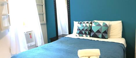 A bedroom with a bed, pillows, a folded towel, a mirror, and a window with curtains.
