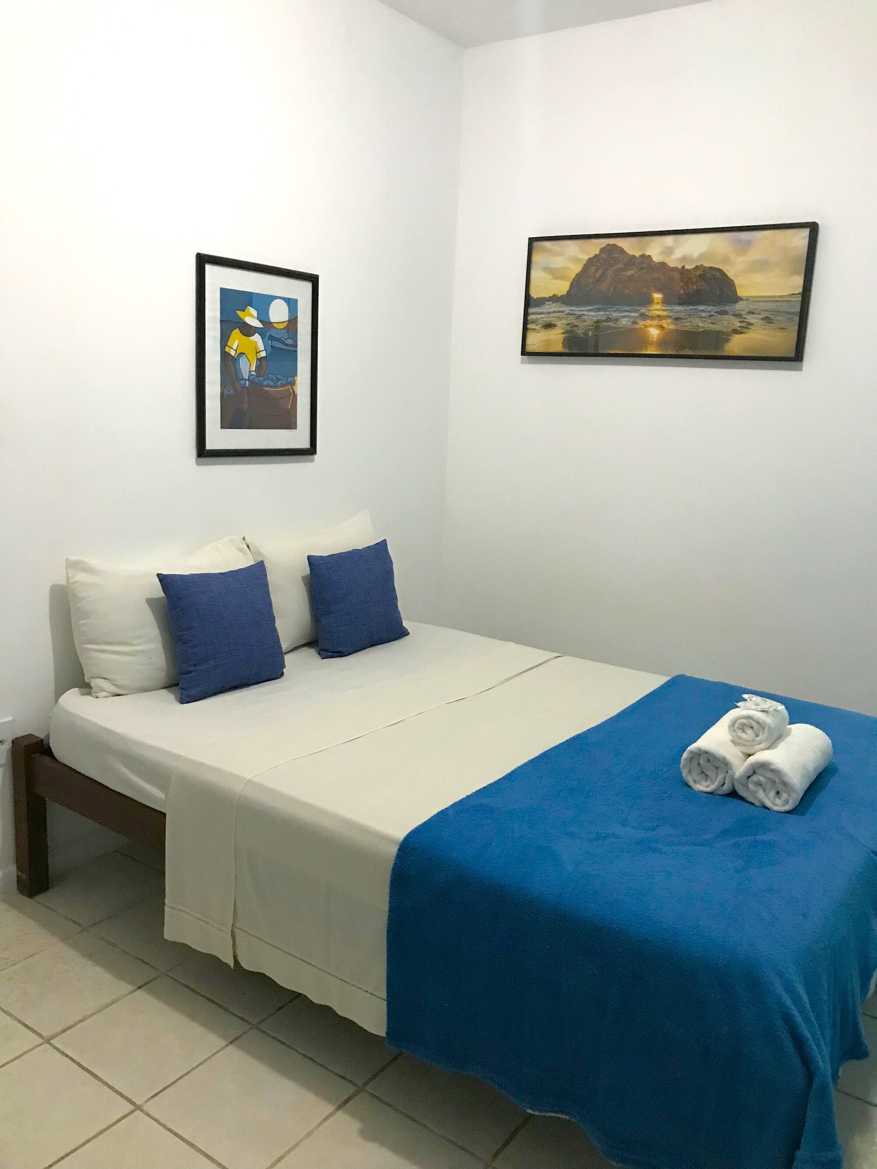 Een netjes opgemaakt bed met een blauwe deken en opgerolde handdoeken, een witte muur met twee ingelijste foto's en een betegelde vloer.