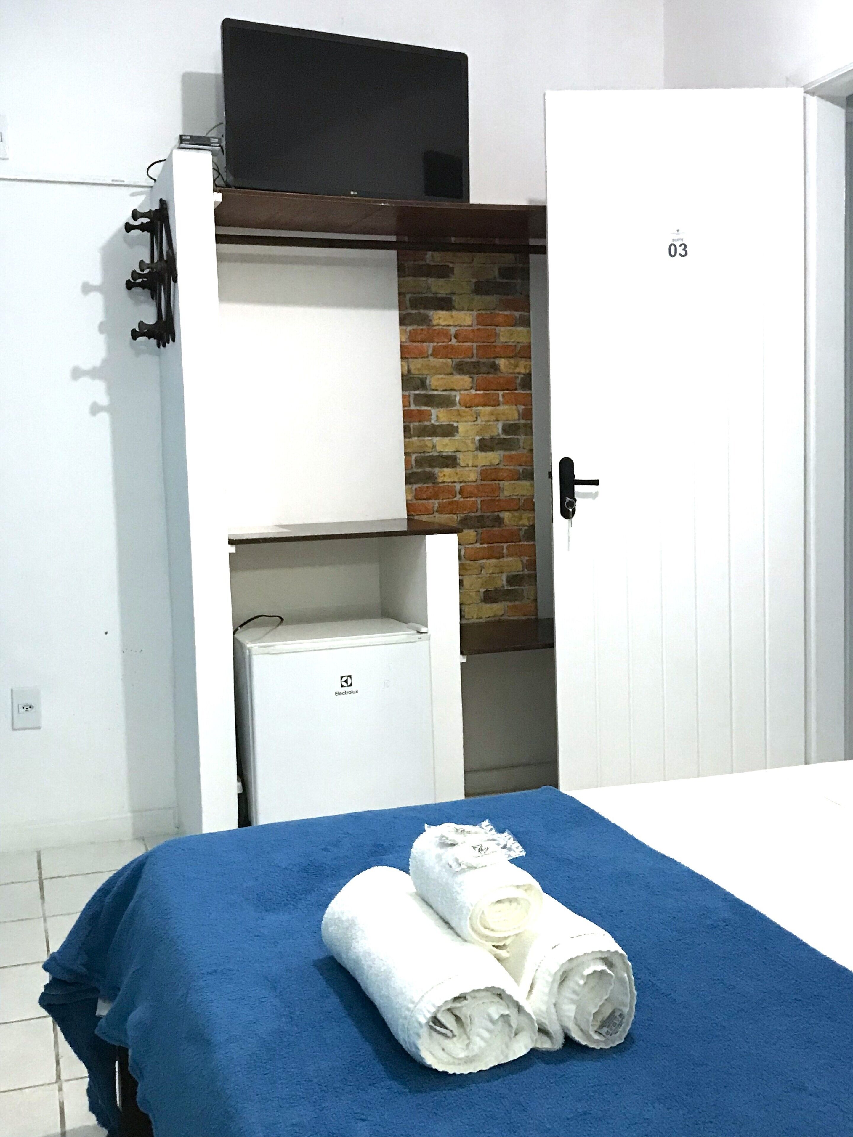 A hotel room with a bed, a small white refrigerator, a brick accent wall, and a television mounted above the bed.