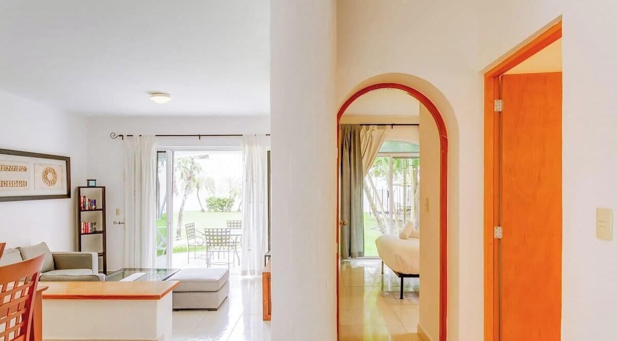 A modern living room with a sofa, a coffee table, and a bookshelf. There is a glass door leading to an outdoor area with a patio table and chairs.