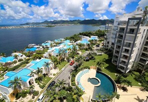 A resort with multiple blue-roofed buildings, a central swimming pool, and a view of the ocean and mountains. - Spacious Garden View Condo (Maho)