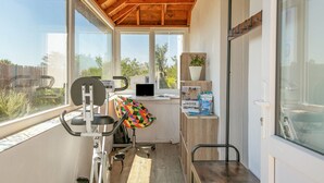 Una habitación iluminada por el sol con techo de madera, una cinta de correr, un sillón de colores, un escritorio con una computadora portátil y una ventana con vistas a zonas verdes y edificios.