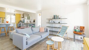 Una sala moderna con un sofá gris, una mesita redonda pequeña y una silla. Se observa una zona de cocina con una barra y taburetes. Hay estanterías con libros y objetos decorativos.