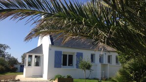 Un edificio blanco con techo gris, un pequeño jardín y una palmera en primer plano.