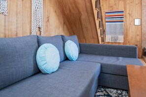 Una sala de estar moderna con un sofá seccional gris, dos almohadas azul claro, techo de madera y un tapiz tejido colorido en la pared.