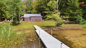 A wooden dock extending into a pond with lily pads, surrounded by trees and a red building with a grey roof.