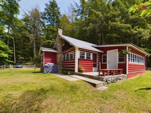 Une cabane en rondins rouge avec une cheminée en pierre, un barbecue couvert et une pancarte de bienvenue.