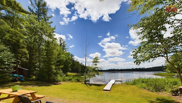 A lakeside picnic area with a wooden table and chairs, a dock extending into the water, and a forested backdrop.