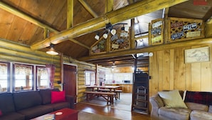 A rustic cabin interior with a wooden ceiling, a dining area, and a living room furnished with sofas and a coffee table.
