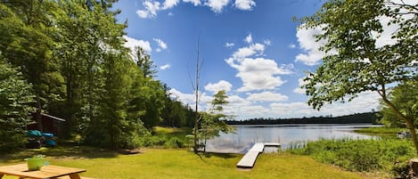 Un área de picnic junto al lago con una mesa y sillas de madera, un muelle que se adentra en el agua y un bosque en el fondo.