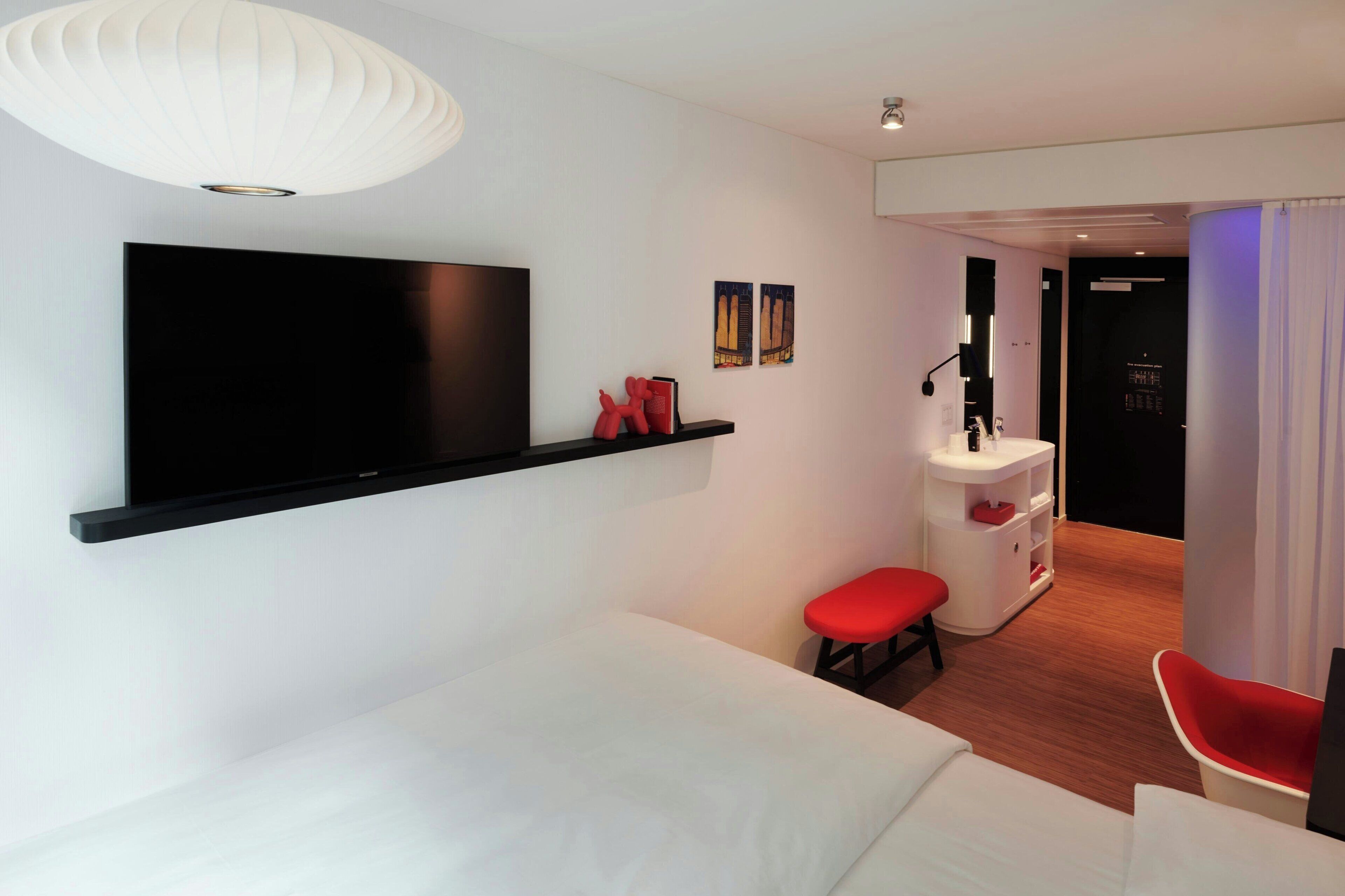 Ein modernes Hotelzimmer mit einem großen Flachbildfernseher an der Wand, einem weißen Sofa, einem roten Sessel, einem kleinen roten Hocker und einem weißen Nachttisch mit Lampe.
