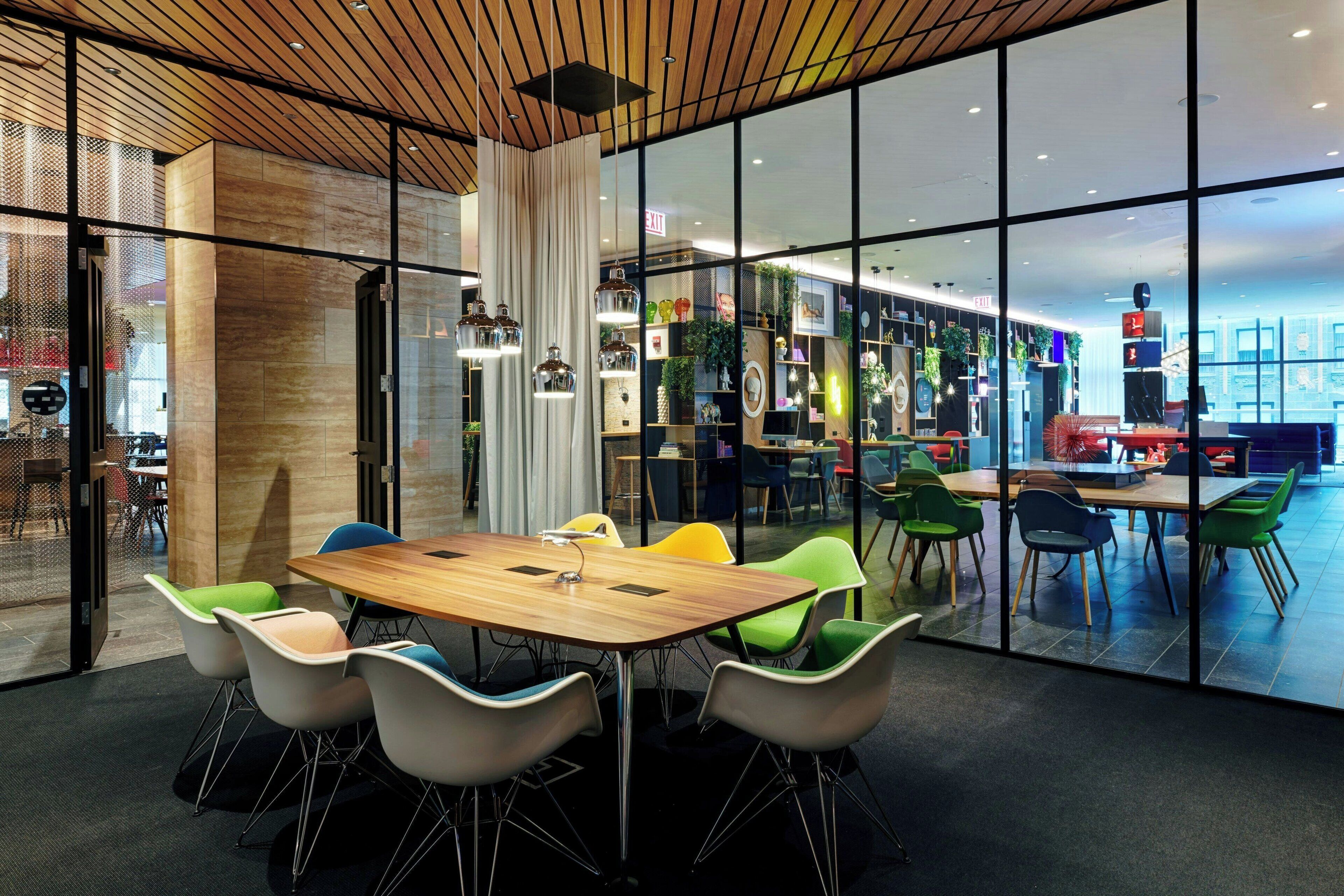 A modern conference room with a wooden table and colorful chairs, large windows, and a view of a restaurant area.