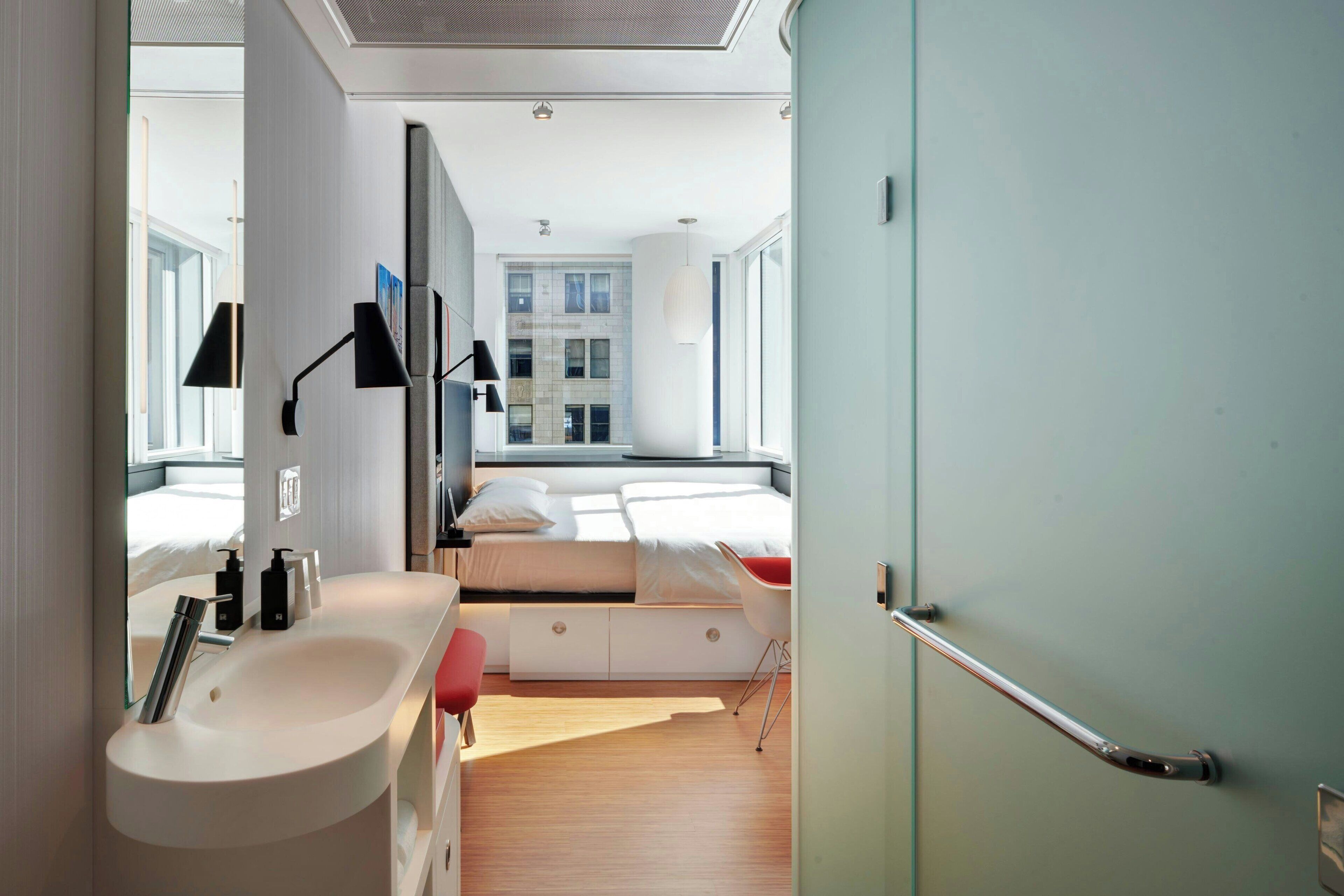 A modern hotel room with a large bed, bedside tables, a built-in bench, and a view of a cityscape.