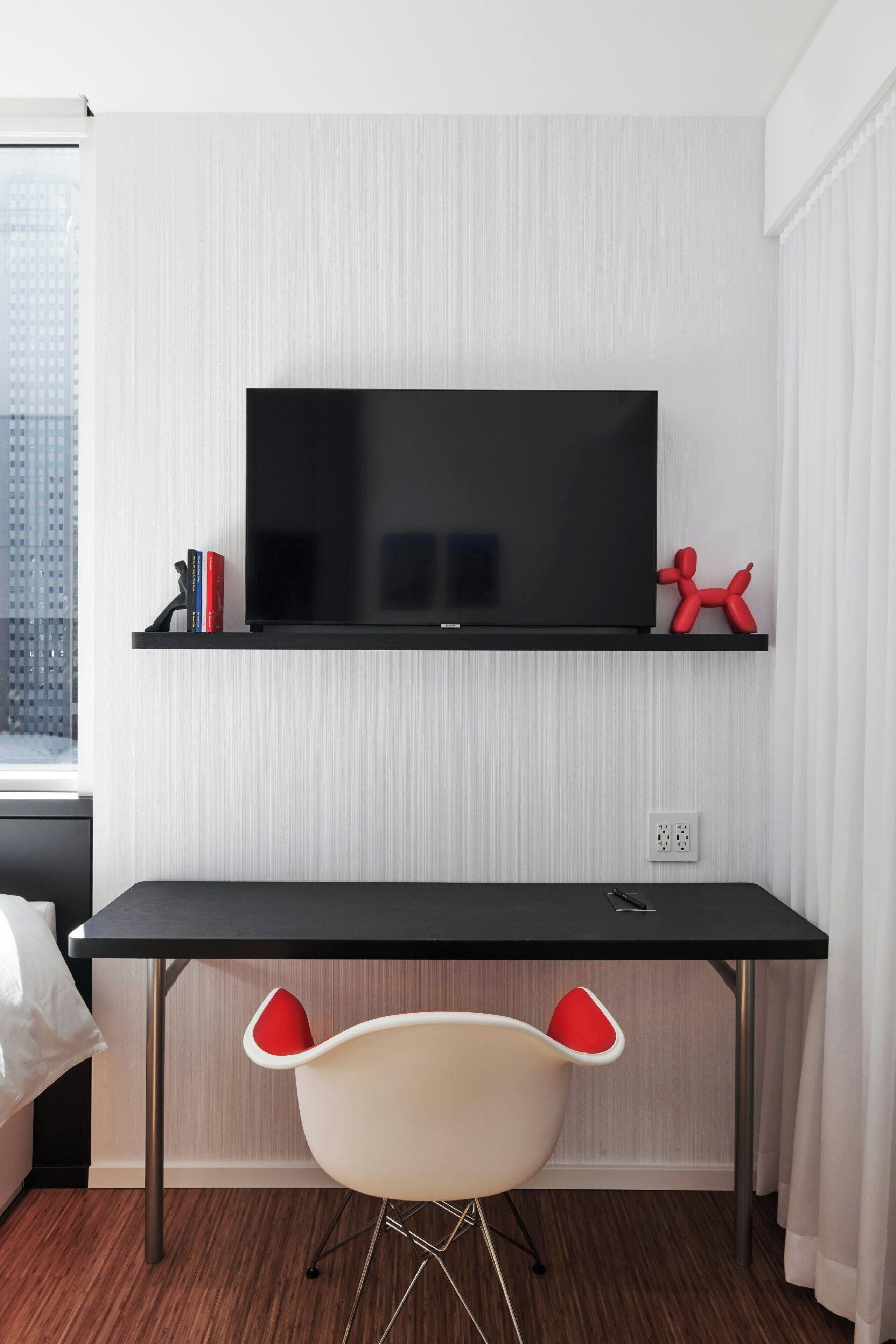 A modern room with a black desk, a television mounted above it, a red balloon sculpture on the shelf, and a white chair with red accents.