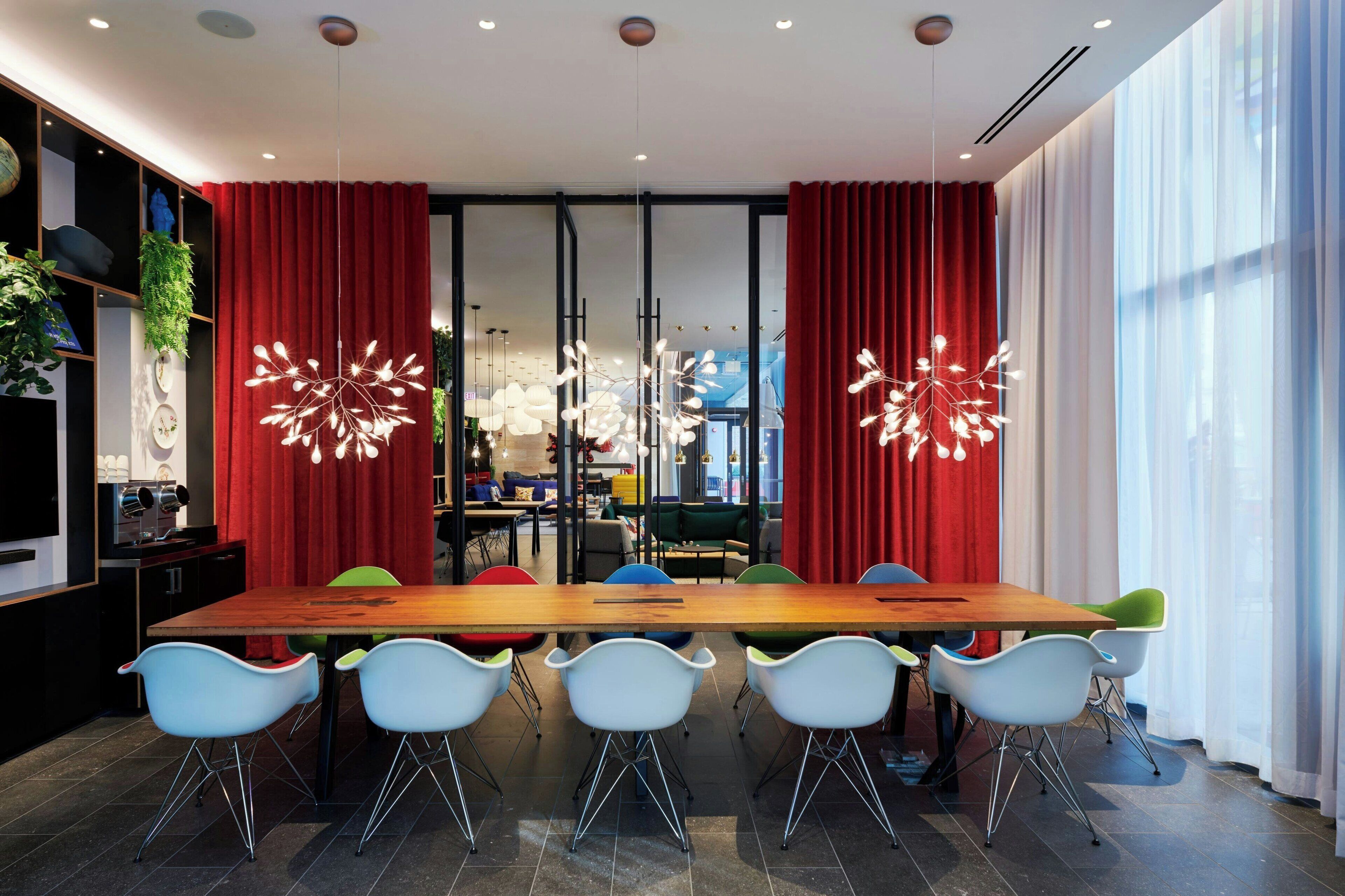 Un comedor moderno con una larga mesa de madera, sillas coloridas y cortinas rojas.