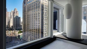 A modern hotel room with a large window offering a city view, a contemporary light fixture, and a clean, minimalist design.