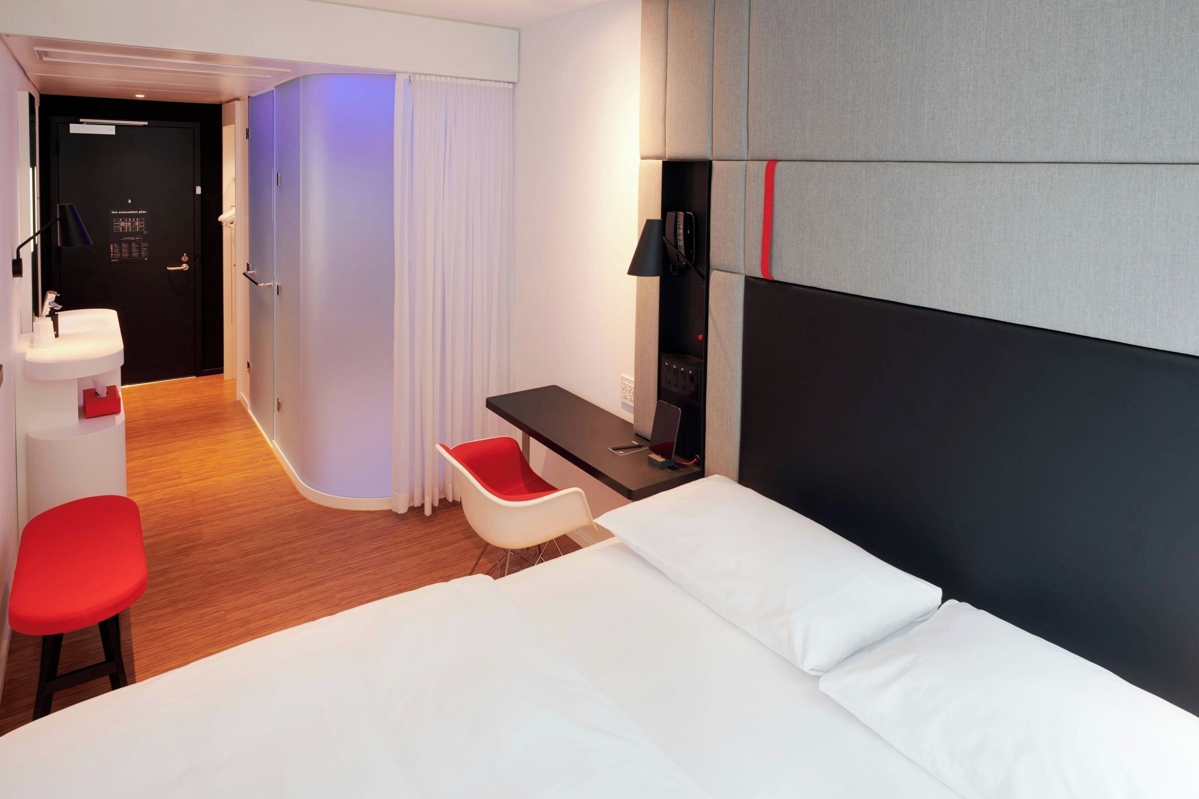Ein modernes Hotelzimmer mit Bett, Schreibtisch, Stuhl, Schreibtischlampe sowie einem Badezimmer mit Waschbecken und Spiegel.