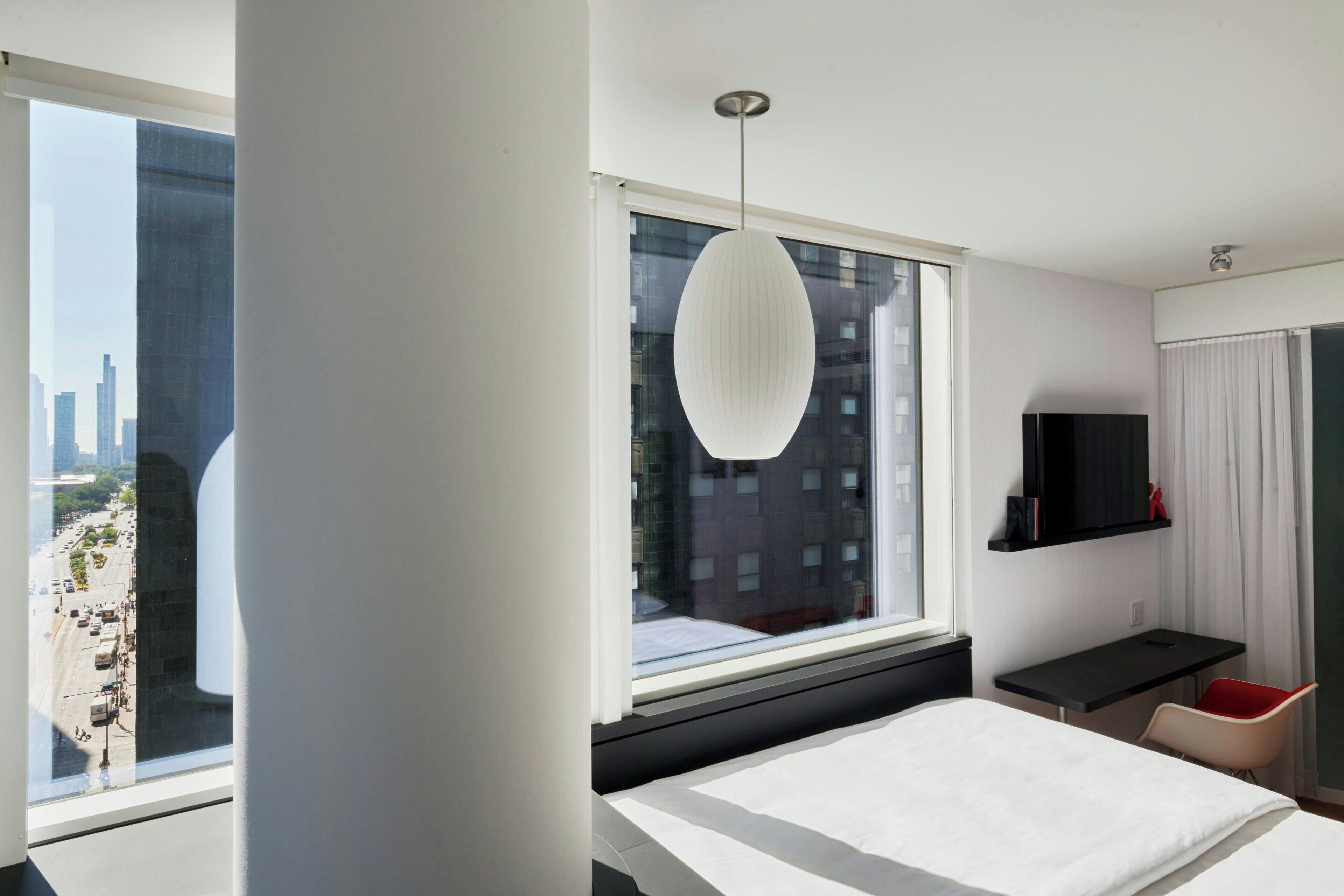 A modern hotel room with a large bed, a desk, a chair, a TV, and a view of the city through a large window.