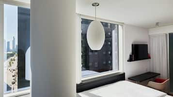A modern hotel room with a large bed, a desk, a chair, a TV, and a view of the city through a large window.