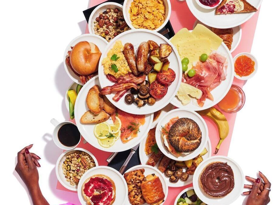 Une variété d’aliments pour le petit-déjeuner sont disposés sur une table, notamment des toasts, des œufs, des saucisses et des fruits.