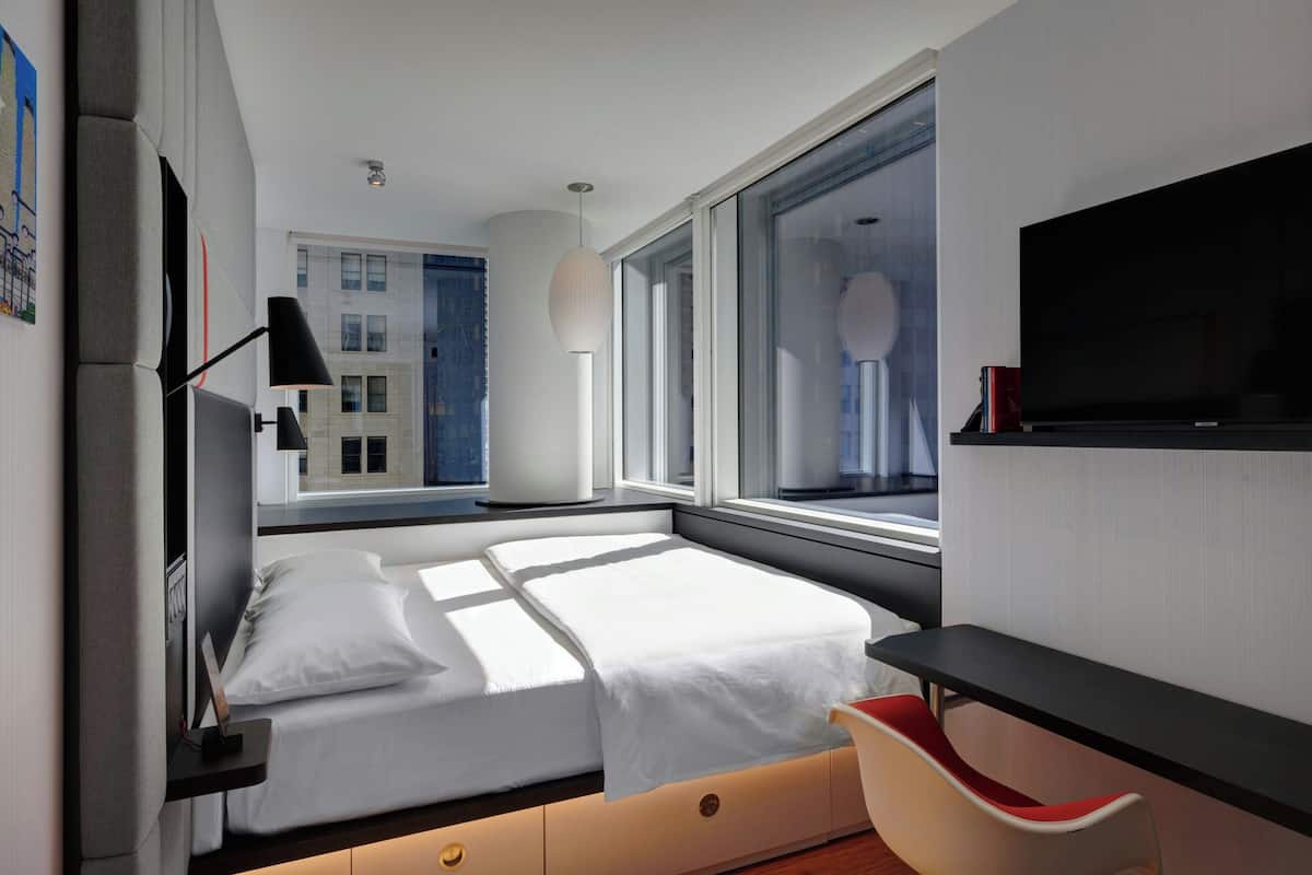 A modern hotel room with a bed, a desk, a chair, a TV, and a window with a city view.