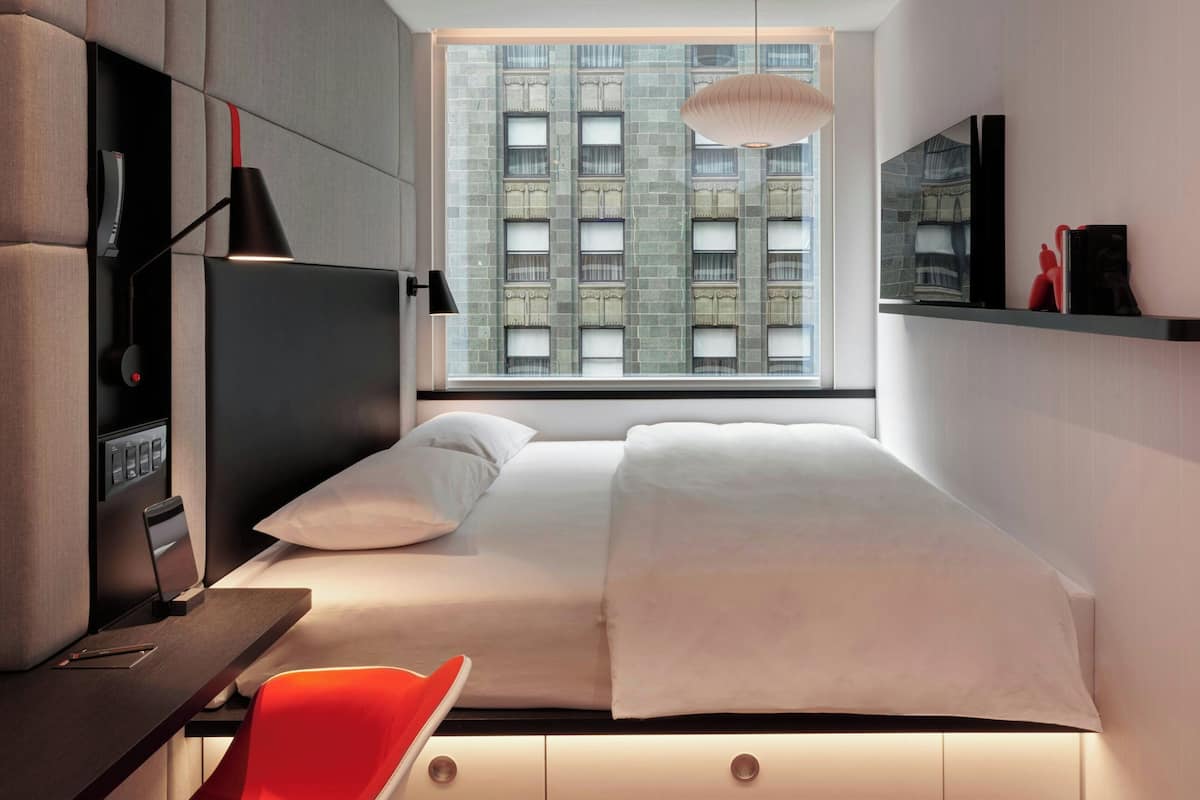 A modern hotel room with a bed, a desk with a chair, a TV, and a window with a city view.