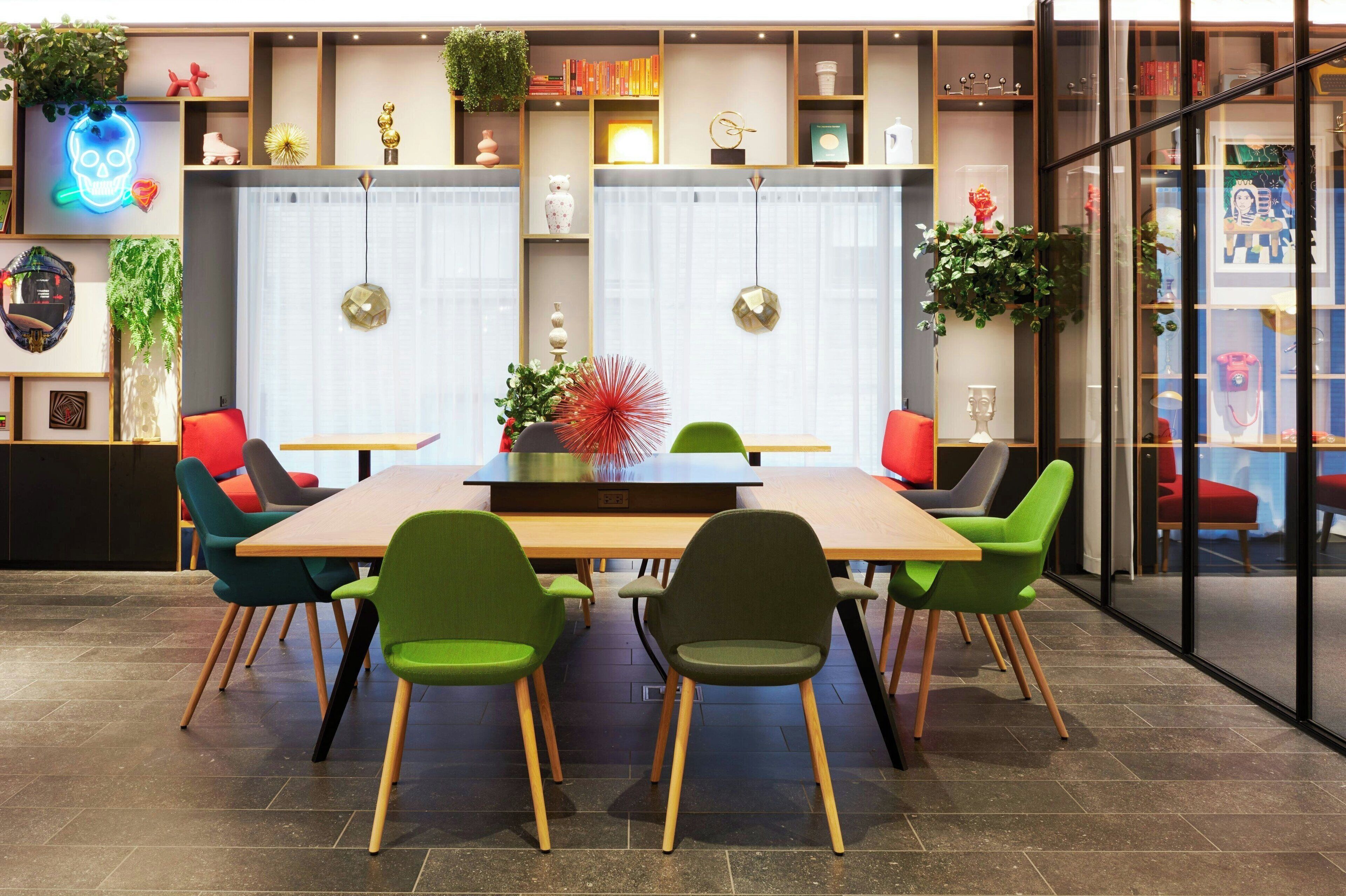 Uma sala de reuniões moderna com mesa de madeira, cadeiras verdes e vermelhas, e prateleiras decorativas com plantas e livros.