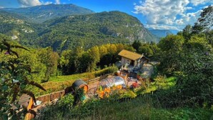 A rustic mountain cabin with a wooden deck, surrounded by lush greenery and a clear blue sky.