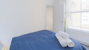 Ein Schlafzimmer mit blauer Bettdecke, weißen Wänden, einer Tür zu einem anderen Raum und einem Fenster mit Jalousien.