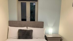 Una cama bien tendida con una toalla blanca enrollada, un plaid gris y un cabecero. Posee una mesita de noche con una lámpara y un cajón. Una ventana con vistas a árboles se encuentra sobre la cama.