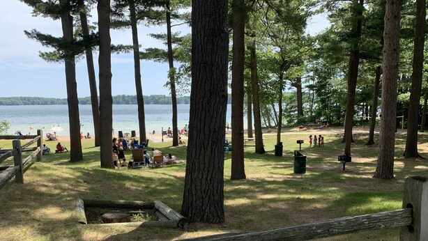 Ein Park mit Holzzaun, Bäumen und einem See, im Hintergrund sind Menschen zu sehen.