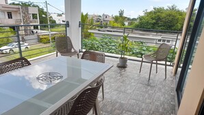 A balcony with a glass table and wicker chairs, overlooking a street and greenery. - GINGER ACCOMMODATION + POOL (Trou aux Biches)