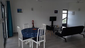 A dining area with a round table, white chairs, a black sofa, and a TV on a stand. - GINGER ACCOMMODATION + POOL (Trou aux Biches)