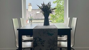A dining area with a table and chairs, a window with a view of buildings and trees, and a plant on the table.