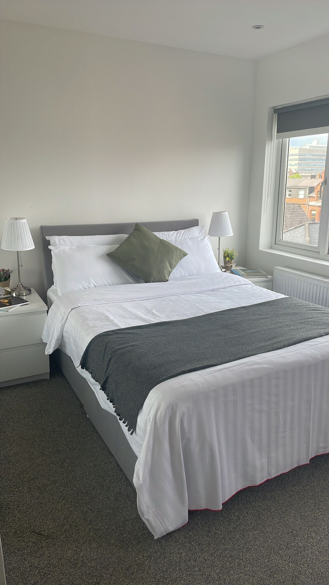 Una cama bien hecha con sábanas blancas y una manta gris, flanqueada por mesitas de noche blancas con lámparas, en una habitación con un gran ventanal que ofrece vistas de edificios.