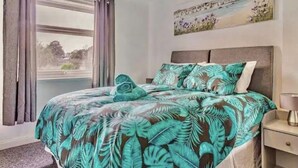 A bedroom with a bed covered in a tropical leaf-patterned bedspread, a nightstand with a lamp, and a painting of a landscape on the wall.