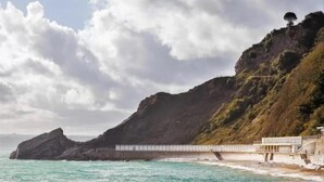 A coastal landscape with a rocky cliff, a bridge, and a building with a balcony overlooking the sea.