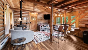 A log cabin interior with a fireplace, sofa, dining area, and staircase. - Small Timbers-riverfront log home, stunning view (Gold Bar)