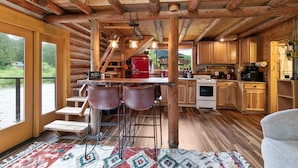 A rustic cabin interior with a wooden staircase, a kitchen area, and a bar with stools. - Small Timbers-riverfront log home, stunning view (Gold Bar)