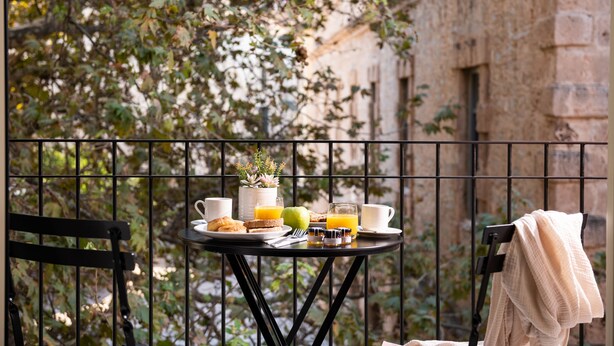 Une table dressée avec des aliments pour le petit-déjeuner sur un balcon, d’où l’on peut admirer un bâtiment en pierre et des arbres.